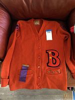 Front view of orange letterman sweater showing large 'B' patch with star pins, purple ribbon and three pins on front pocket area, and the button front closure with ribbed cuffs.