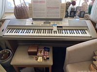 Full view of Yamaha DGX-500 keyboard with vintage song books on music stand, keyboard stool below with small personal items on it, beige chair to side
