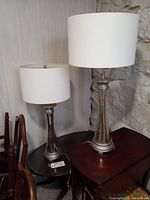 Two electric table lamps with curved mercury glass-like bases and round white fabric shades sitting on wooden tables against a stone and paneled wall background.