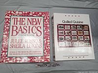 Photo showing both cookbooks side by side on a gray surface. Quilted Quisine on the right with white cover and quilt pattern. The New Basics Cookbook on the left with red and white cover and visible text.