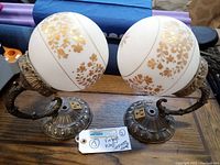 Two round-glass gold leaf decorated brass wall sconces with curved arms and ornate base, viewed from above and front.