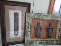Two framed African mask shadow box art pieces displayed side by side, showing one frame with two metallic masks on burlap and one frame with single wooden mask.