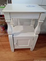 Front view of an off-white cream side table showing top surface, shelf, turned legs, and closed bottom cabinet door with knob.