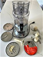 Photo showing the food processor with stainless steel base, clear plastic bowl and lid, power cord, three slicing/shredding discs and attachments arranged around it.