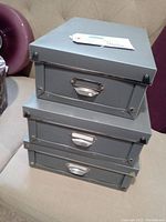 Three stacked grey storage boxes with lids and metal reinforcements, showing condition and design details.