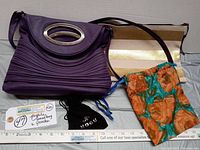 Photo showing the purple handbag with fold-over flap, silver-tone oval handle detail, and four drawstring pouches in front.