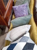 Five decorative toss pillows arranged on a mustard yellow couch including a purple floral embroidered pillow, two green-grey textured pillows, a light grey plain pillow, and a geometric patterned pillow.