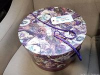 Top view of vintage Bradlees hat box showing purple floral collage lid and braided cord handle with retail tag attached.