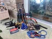 Photo showing collection of hand tools, power tools including chainsaw, saws, buffers, hedge trimmer, tools and jumper cables on a carpeted surface by large window.