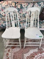 Two white painted wooden chairs with spindle backs and visible scratches and cracks, placed on a floral couch and patterned rug.
