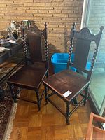 Pair of antique wooden dining chairs shown side by side on a wood floor in front of brick wall. Chairs have carved crest, spiral legs and uprights.