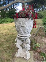 Front view of concrete planter with red flowering plant inside, showing lion-themed base and classical relief detail around planter bowl.