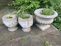 Three concrete planters on stone surface next to green foliage; two smaller with Greek key pattern and one large with rounded rim