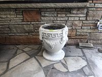 Front view of concrete urn showing Greek key pattern and grapevine decoration with soil inside.