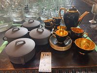 Photo showing four gray Dansk lidded bowls, each with a handle on the lid, placed on the left side of the table. To the right are black and gold ceramic pieces including cups and a pot. The lot tag is placed near the bowls.