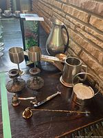 Overview of 12 items including metal pitcher, stein, goblets, coasters, solar light, candle snuffer, candle holders, cutlery, and strainer on dark table in front of brick wall.