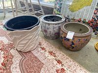 Four planters displayed on a patterned carpet: three ceramic planters of different decorative styles and one black resin flowerpot insert.