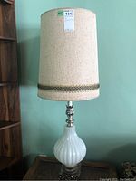 Full view of one lamp showing beige fabric shade with trim, opaque white ribbed glass base, and metal neck accents.