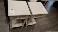 Two square wooden end tables with mirrored decorative drawers and a lower shelf, painted beige, showing wear on the surfaces.