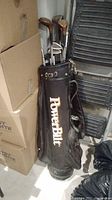 Photo of black PowerBilt golf bag filled with golf clubs, showing wear and environment.