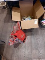 Box with assorted items including cleaning product packet, plastic bags, and small items inside an open cardboard box on floor.