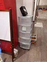 Five drawer clear plastic storage unit positioned against wall with Zeus cleaning kit box on top, kitchen visible in background, floor dog feeding station adjacent.