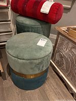 Photo of two round covered ottomans stacked, one green and blue with gold band, one solid green, with a red bolster cushion on top.