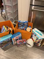 Photo showing multiple boxed puzzles, construction kits, and wooden toys arranged on a floor near storage bins.