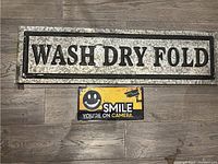 Photo showing both metal signs on wood floor: the large 'WASH DRY FOLD' sign and the smaller 'SMILE YOU'RE ON CAMERA' sign.