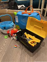 Full lot showing blue Trunki suitcase, yellow and black Stanley toy toolset, blue plush whale basket, and two plush dolls on the floor.