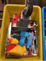 Yellow rubber duck, blue Thomas the Tank Engine train, grey and red toy structure, plastic Pixar character dog toy in yellow bin