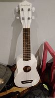 Front full view of white ukulele leaning against a wall showing body, fretboard, and headstock.