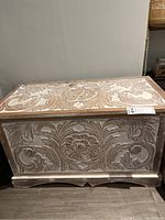 Front view of an off-white carved wooden chest showing floral and leaf motifs on the front panel and top.