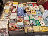 Photo of a large assortment of cookbooks and kitchen guides laid out on beds, showing a variety of titles, cover art, and formats.