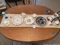 Overview of assorted fine china plates, cups, saucers, decorative bell, candlestick holders, and blue stemmed glass arranged on dining table.