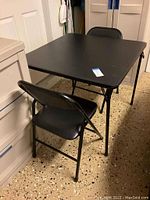 Black square folding card table with two black folding chairs set around it, showing table surface and chair details.