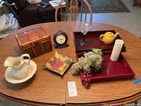 Table top displaying wooden serving trays, treasure chest, clock, ceramic pitcher and basin, decorative dish, faux grapes, glass candle holder, white candle, and a yellow ceramic teapot.