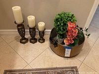 Photo showing three decorative brass candlesticks of different heights with white LED candles next to a large brass kettle with artificial floral arrangement.