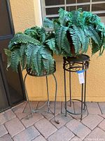 Two faux fern plants in decorative metal stands displayed outdoors against a stucco wall. Left plant and stand shorter than right.