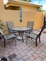 Full view of the patio furniture set on stone pavers including four chairs, glass top table, and umbrella closed inside table's umbrella hole.