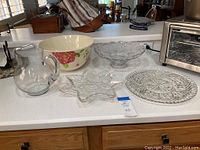 Kitchen countertop displaying all items in the lot including glass pitcher, floral ceramic bowl, cut glass pedestal bowl, star-shaped glass dish, and round cut glass platter.