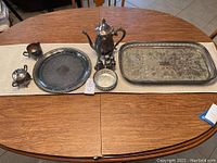 Full view of the silverplate set arranged on a wooden table on a beige runner. Items include teapot, sugar bowl, creamer, salt and pepper shakers, small bowl, round platter, oblong tray, and small bell.