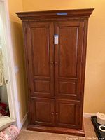 Full frontal view of the wood TV armoire standing against a beige wall. Shows two upper doors and two lower drawers with round wood knob handles. Medium brown finish with visible wood grain and crown molding top.