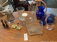 Photo showing all items in the lot arranged on a dining table including the blue glass vases, clear glass vases, candle sticks, flower frog, and wire rack.