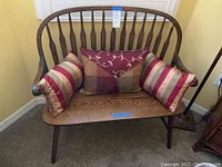 Full view of the vintage wooden bench with three decorative pillows placed on the seat against a beige wall and carpeted floor.