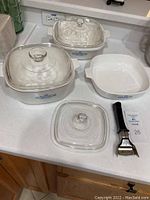 Photo showing the full vintage Corning Ware set of three white casserole dishes with cornflower blue flower designs, three glass lids with knobs, and the detachable black-handled metal holder.