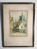 Framed serigraph print showing an old street scene with cottages and a church tower, signed and titled in pencil below image.