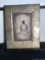 Front view of the original metal art piece, showing the textured metal frame and the sketched figure seated with back facing viewer.