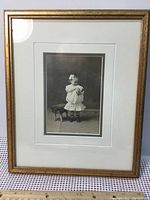 Frontal photo of the antique baby photograph showing the girl in a white dress, seated, surrounded by a white mat and gold-colored frame.