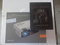 White Apeman LED50 Digital Projector placed on top of its box alongside the Game of Thrones The Complete First Season DVD set with Iron Throne cover art.